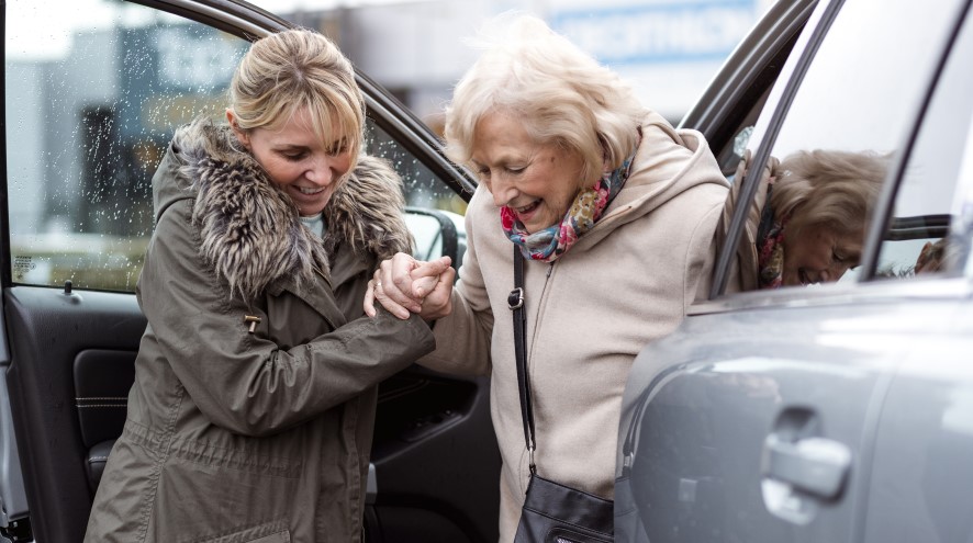 Middle-aged woman helping a senior woman out of her car.