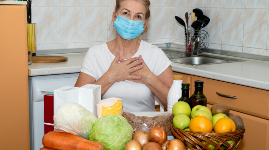 Image of a woman holding her heart in thanks, wearing a mask and surrounded by fresh vegetables and fruit